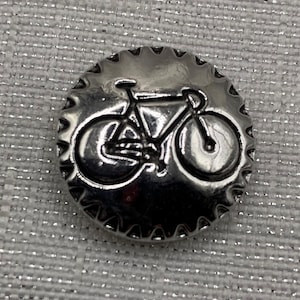 May include: A silver-colored, round button with a bicycle design in the center. The button has a scalloped edge. The bicycle is a detailed outline, showing the frame, wheels, and other components. The button is likely a decorative element.