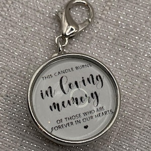 May include: A round, silver-toned charm with a lobster clasp. The charm features the text "This candle burns in loving memory of those who are forever in our hearts" in black script on a gray background.