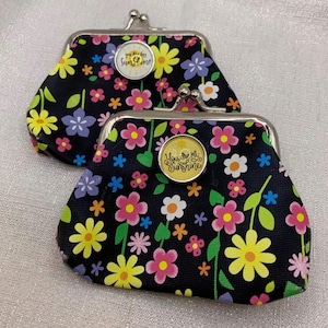 May include: Two coin purses with a black background featuring a colorful floral pattern of yellow, pink, and purple flowers. Each purse has a silver metal kiss-lock clasp and a circular center emblem with the text You are my sunshine.