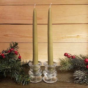 May include: Two olive green taper candles in clear glass candle holders. The candle holders have a decorative, layered design. The candles are surrounded by evergreen branches with red berries, set against a wooden background.