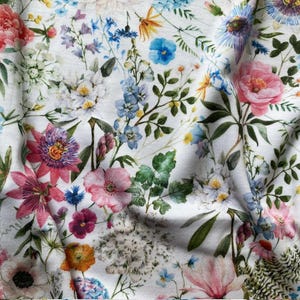 Op de afbeelding: Witte stof met een aquarel bloemenpatroon. Het ontwerp toont een verscheidenheid aan bloemen in roze, blauwe, paarse en gele tinten, met groene bladeren. De stof heeft een zachte, vloeiende uitstraling, wat suggereert dat hij licht van gewicht is en geschikt is voor kleding of woondecoratie.