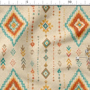 May include: A beige fabric featuring a repeating geometric pattern of diamonds and vertical lines in shades of orange, teal, and yellow. A metal ruler is visible along the top and left edges, and several silver safety pins are attached to the fabric.