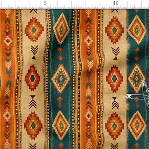 Puede incluir: Una tela con un patrón vertical repetitivo de diamantes y formas geométricas en verde azulado, naranja y crema. Una regla metálica en los bordes superior e izquierdo muestra una escala en centímetros, y un imperdible plateado descansa sobre la tela.