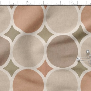 Könnte beinhalten: Ein Stoff mit einem sich wiederholenden geometrischen Muster aus großen Kreisen in Pfirsich-, Beige- und Brauntönen, durchsetzt mit kleinen olivgrünen Rauten. Eine Sicherheitsnadel aus Metall ist am Stoff befestigt, der neben einem Lineal liegt.