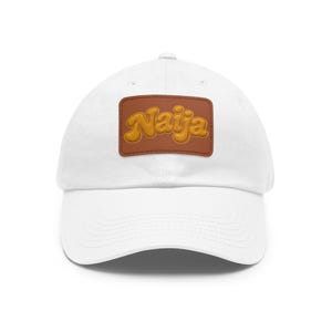 Puede incluir: Una gorra de béisbol blanca con un parche rectangular de cuero marrón centrado en la parte delantera. El parche muestra la palabra Naija en letras doradas, redondeadas y de estilo retro, con costuras visibles en los bordes.