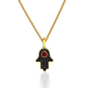 May include: A gold chain necklace with a hamsa hand pendant. The pendant is covered in black cubic zirconia stones with a single red cubic zirconia stone in the center.