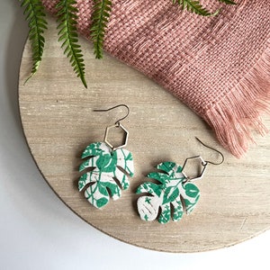 May include: A pair of green and white leaf-shaped earrings with a silver hexagonal charm. The earrings are made of cork and have a floral pattern.