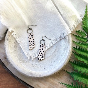May include: A pair of dangle earrings with a black and white spotted pattern. The earrings are made of leather and have a hexagonal metal finding.
