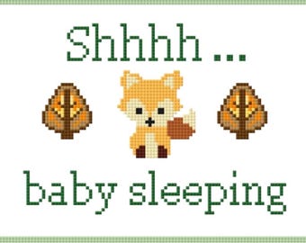 May include: Cross-stitch design featuring a cute orange fox with a white belly, flanked by two stylized trees. The text "Shhhh... baby sleeping" is stitched in green, set against a white background with a green border.