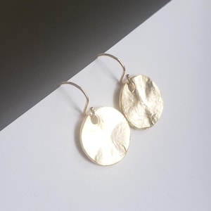 May include: A pair of gold-toned drop earrings featuring circular, hammered metal discs. Each disc has an irregular, textured surface and is suspended from a simple curved ear wire hook, creating a reflective and metallic appearance.