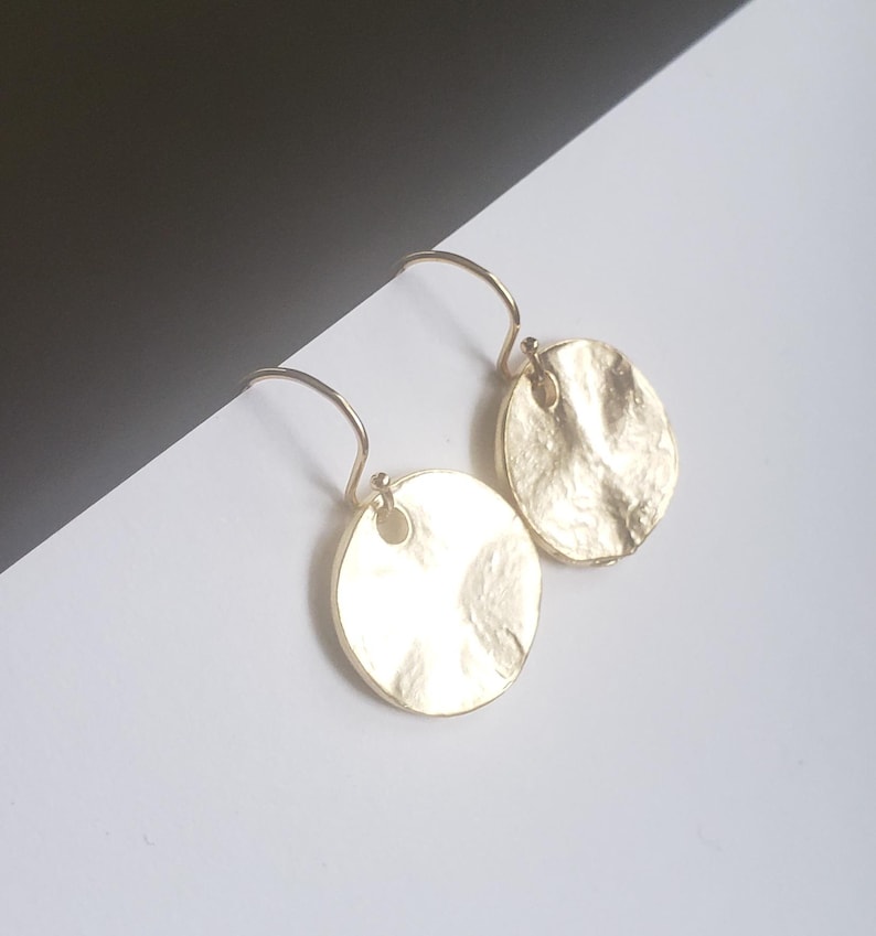May include: A pair of gold-toned drop earrings featuring circular, hammered metal discs. Each disc has an irregular, textured surface and is suspended from a simple curved ear wire hook, creating a reflective and metallic appearance.