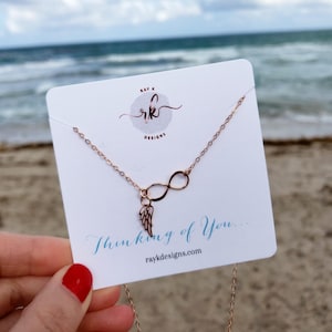 May include: Rose gold infinity necklace with a small angel wing charm. The necklace is displayed on a white card with the text "Thinking of You..." and the website address "raykdesigns.com".