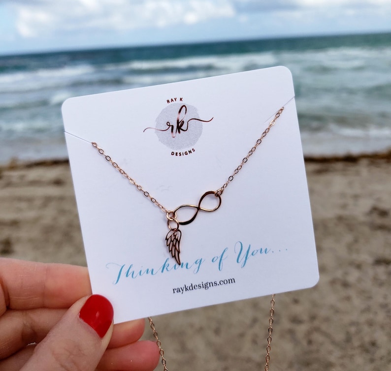 Pu&ograve; includere: Collana infinita in oro rosa con un piccolo ciondolo ad ala d'angelo. La collana &egrave; esposta su una carta bianca con il testo "Thinking of You..." e l'indirizzo del sito web "raykdesigns.com".