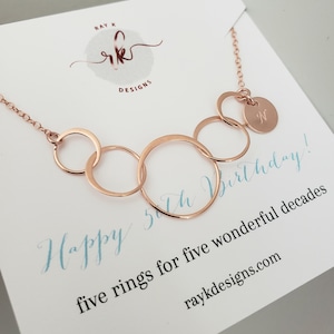 Puede incluir: Collar de oro rosa con cinco anillos interconectados y un peque&ntilde;o colgante con la letra "N". El collar se muestra en una tarjeta blanca con el texto "Happy 5th Birthday!" y "five rings for five wonderful decades".