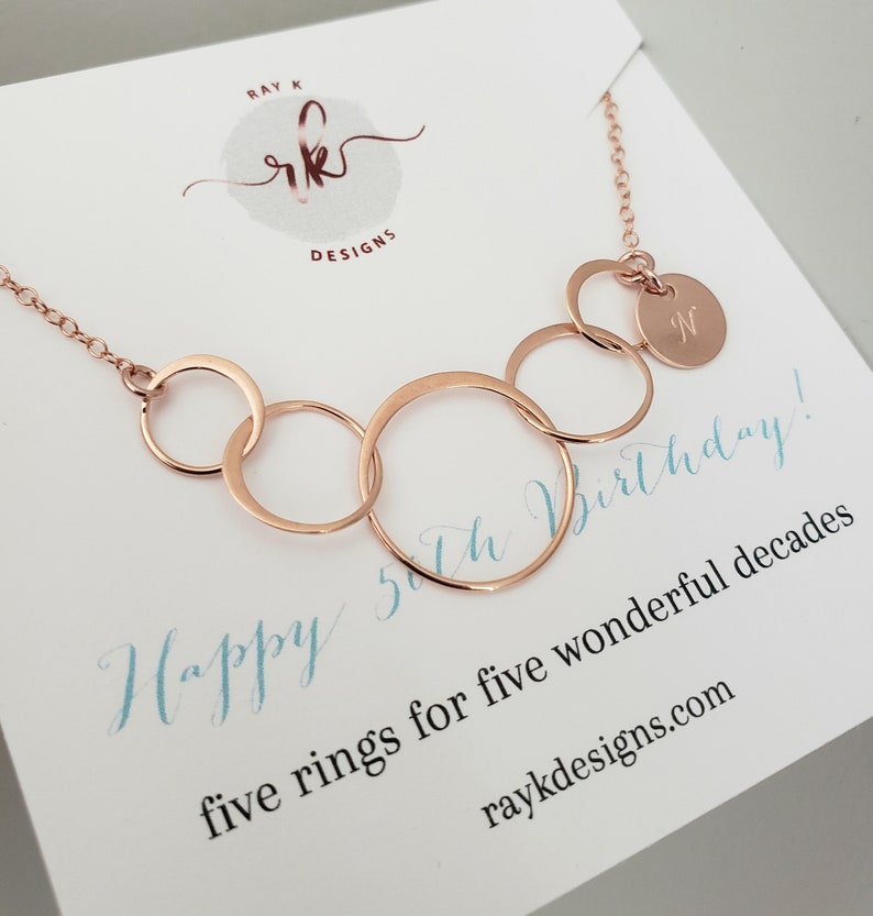 Puede incluir: Collar de oro rosa con cinco anillos interconectados y un peque&ntilde;o colgante con la letra "N". El collar se muestra en una tarjeta blanca con el texto "Happy 5th Birthday!" y "five rings for five wonderful decades".