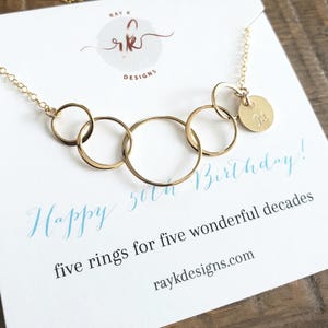 May include: A gold-colored necklace with five interlocking rings and a small, round charm engraved with the letter "M". The necklace is displayed on a white card with the text "Happy 50th Birthday!" and "five rings for five wonderful decades."