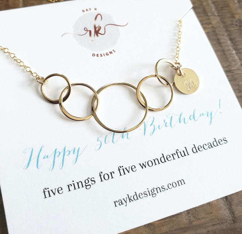 Puede incluir: Un collar dorado con cinco anillos entrelazados y un peque&ntilde;o dije redondo grabado con la letra "M". El collar se exhibe en una tarjeta blanca con el texto "Happy 50th Birthday!" y "five rings for five wonderful decades".
