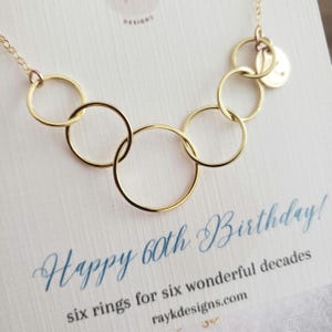 May include: A gold necklace with six interconnected rings and a small disc charm. The necklace is displayed on a card with the text "Happy 60th Birthday!" and "six rings for six wonderful decades."