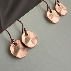 May include: A pair of rose gold-toned drop earrings featuring small, circular, hammered metal discs suspended from curved ear wires. The discs have an irregular, textured surface with a soft metallic sheen and a single hole at the top.