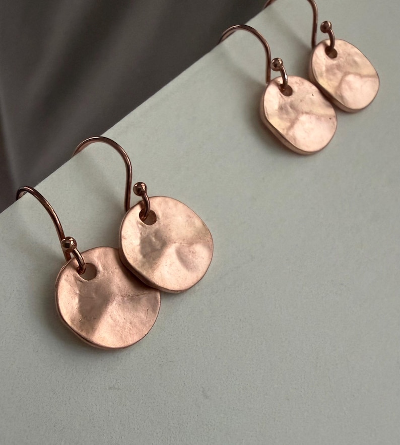 May include: A pair of rose gold-toned drop earrings featuring small, circular, hammered metal discs suspended from curved ear wires. The discs have an irregular, textured surface with a soft metallic sheen and a single hole at the top.