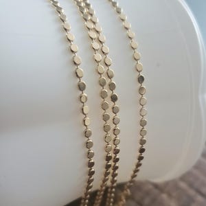 May include: Close-up of several delicate gold necklaces. Each necklace features a chain of small, flat, circular discs. The necklaces are draped over a white surface, showcasing their design and shine. The warm gold color contrasts with the white background.