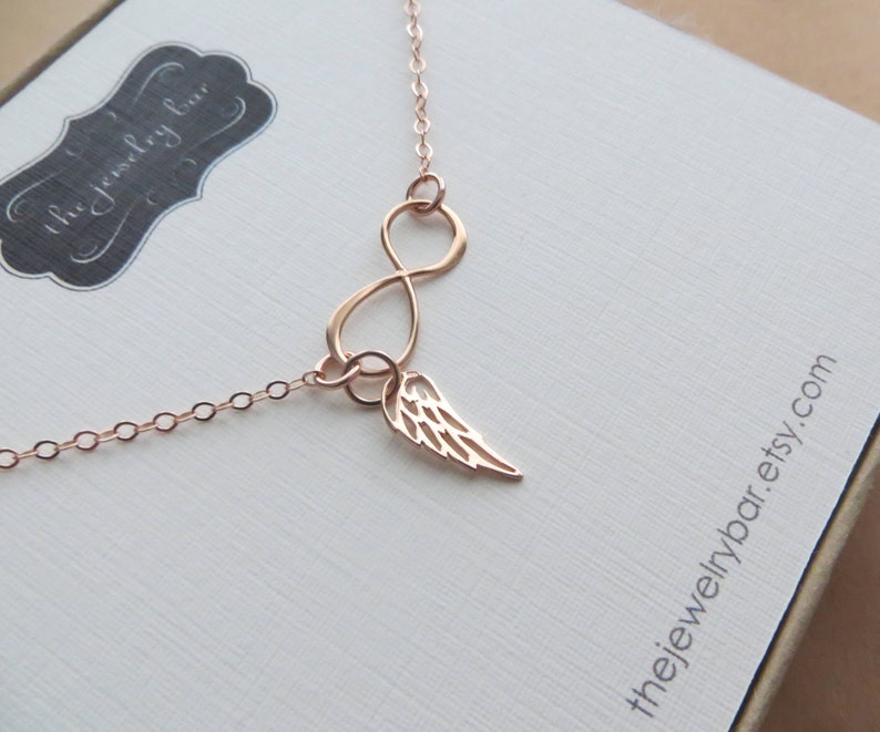 Pu&ograve; includere: Collana infinita in oro rosa con un delicato ciondolo ad ala d'angelo. La collana &egrave; su uno sfondo bianco con il testo "thejewelrybar.etsy.com".