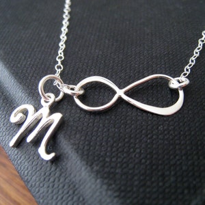 Puede incluir: Un collar plateado con un símbolo de infinito y un colgante con la letra "M" en cursiva. La cadena es fina y el collar se muestra sobre un fondo oscuro.
