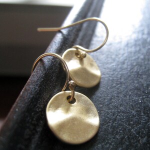 May include: A pair of gold-toned drop earrings featuring small, circular, hammered metal discs suspended from curved ear wires. The discs have a matte, textured finish and are attached to the wires with a small loop, creating a simple, dangling design.