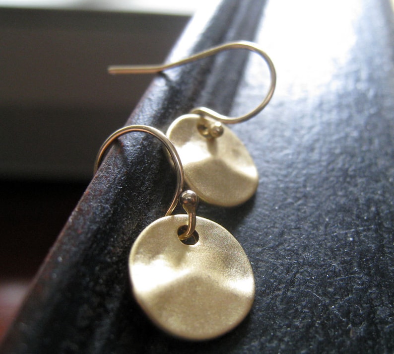 May include: A pair of gold-toned drop earrings featuring small, circular, hammered metal discs suspended from curved ear wires. The discs have a matte, textured finish and are attached to the wires with a small loop, creating a simple, dangling design.