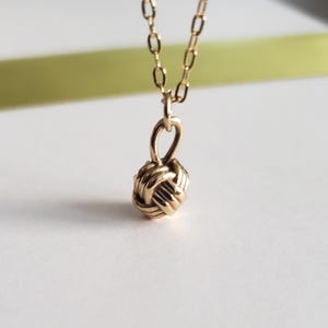 May include: A delicate gold-colored necklace featuring a woven knot pendant. The pendant is intricately designed with interwoven strands, suspended from a fine chain. The necklace is displayed against a soft, blurred background, highlighting the jewelry's elegant simplicity.
