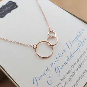 May include: A rose gold necklace with three interlocking circles. The necklace is on a white background with the text "Grandmother Daughter & Granddaughter" written in blue cursive.