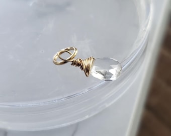 Puede incluir: Un colgante de cristal de cuarzo transparente envuelto en alambre dorado. El colgante tiene forma de lágrima y tiene un bucle de alambre dorado en la parte superior para sujetarlo a una cadena.