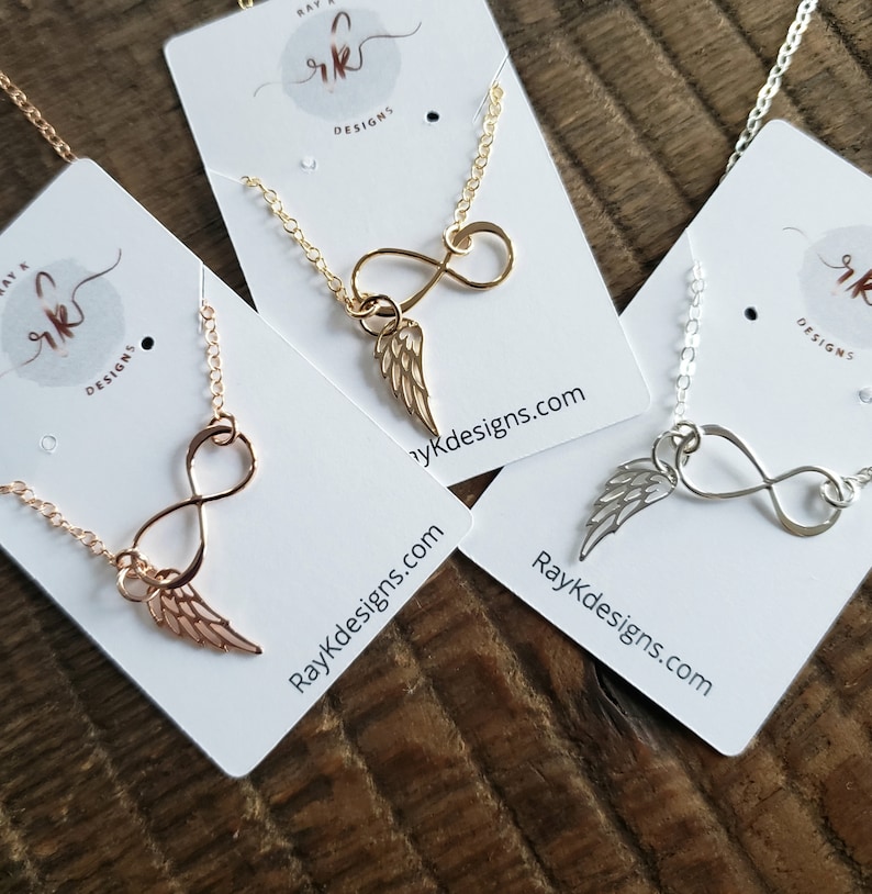 Pu&ograve; includere: Tre collane con simboli dell'infinito e ciondoli ad ala d'angelo. Le collane sono su carte bianche con il testo "Ray K Designs" e l'indirizzo del sito web "RayKDesigns.com".
