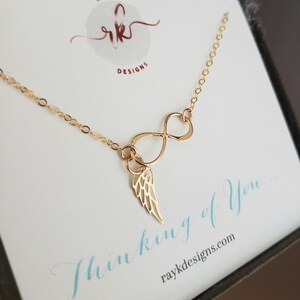 Pu&ograve; includere: Collana infinita in oro con un piccolo ciondolo ad ala d'angelo. La collana &egrave; esposta su una carta bianca con il testo "Thinking of You..." e l'indirizzo del sito web "raykdesigns.com".