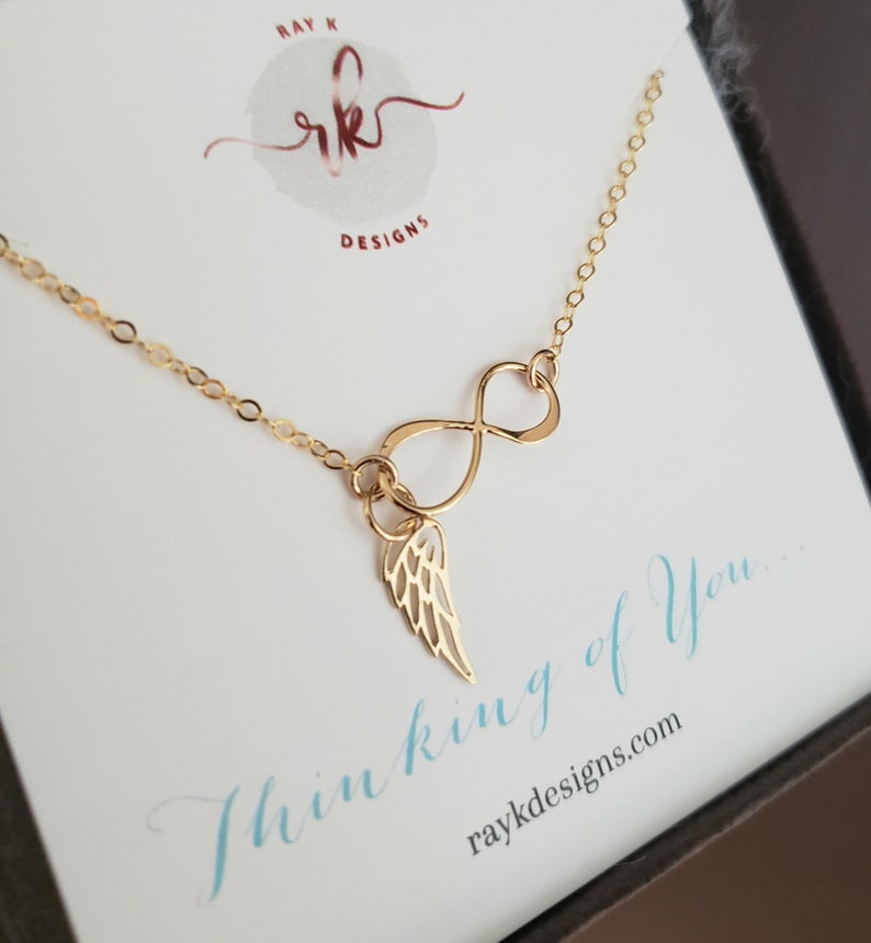 Pu&ograve; includere: Collana infinita in oro con un piccolo ciondolo ad ala d'angelo. La collana &egrave; esposta su una carta bianca con il testo "Thinking of You..." e l'indirizzo del sito web "raykdesigns.com".