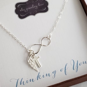 May include: Silver necklace with an infinity symbol, a cross charm, and an angel wing charm. The necklace is displayed in a box with the text "Thinking of You".