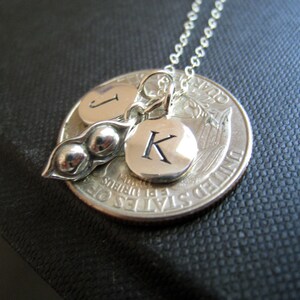 May include: Silver necklace with a charm of two pea pods and two initial charms. The initial charms are engraved with the letters "J" and "K". The necklace is hanging from a silver chain and is attached to a silver quarter.