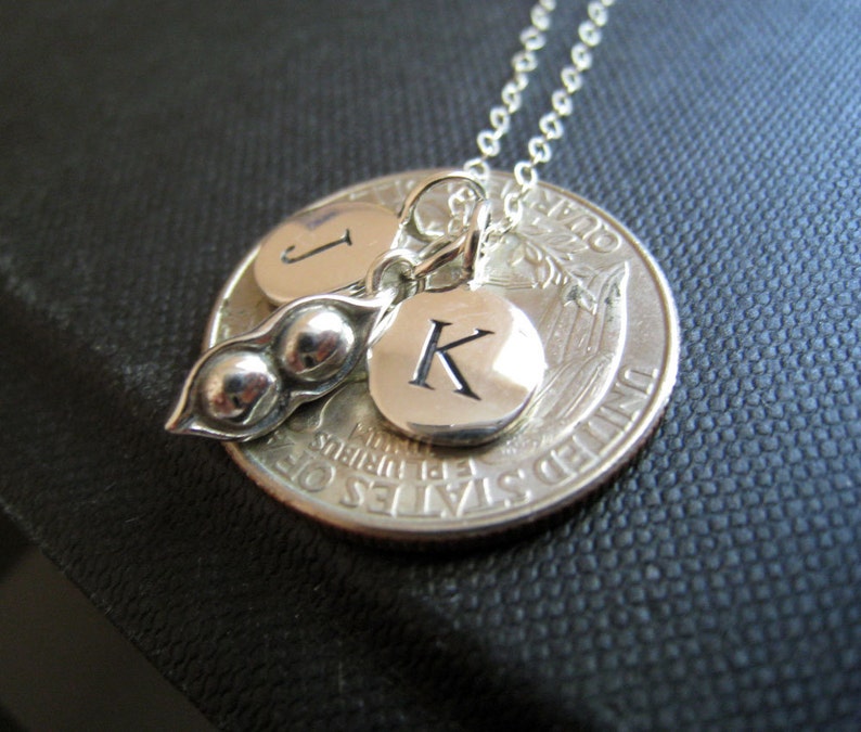 May include: Silver necklace with a charm of two pea pods and two initial charms. The initial charms are engraved with the letters "J" and "K". The necklace is hanging from a silver chain and is attached to a silver quarter.