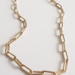 May include: Gold-colored paperclip chain necklace. The necklace features a series of elongated rectangular links, creating a modern and minimalist design. The chain is displayed on a white surface.