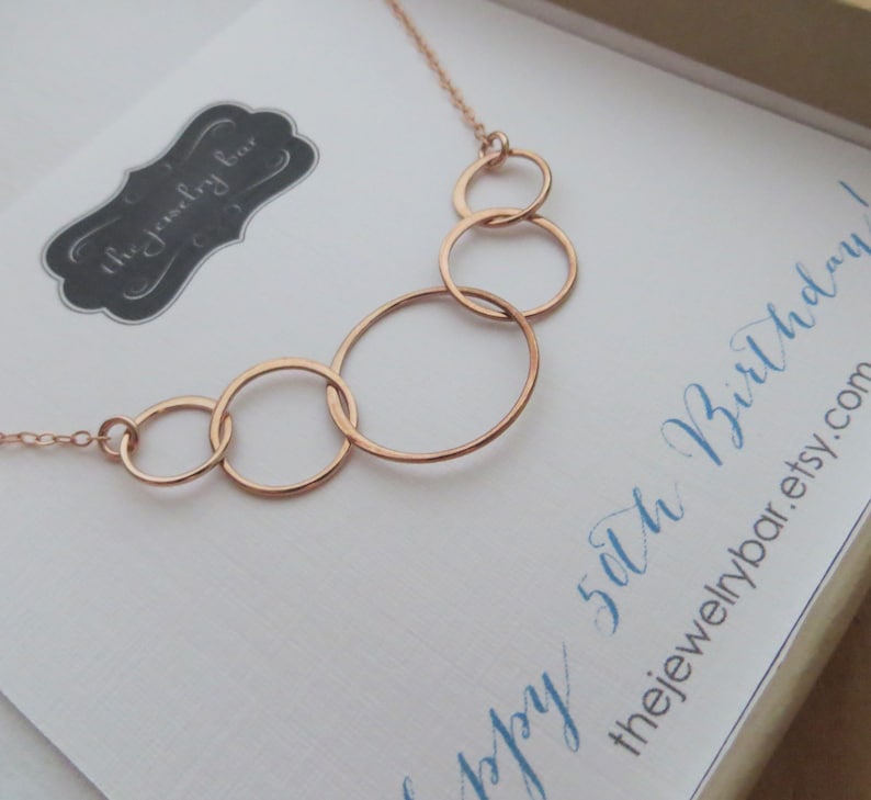 May include: A rose gold necklace with five interlocking circles. The necklace is displayed on a white card with the text "Happy 50th Birthday! thejewelrybar.etsy.com"