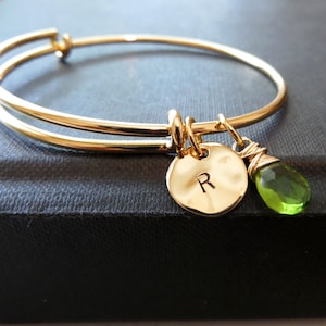 May include: Gold bangle bracelet with a charm featuring the letter "R" and a green gemstone.