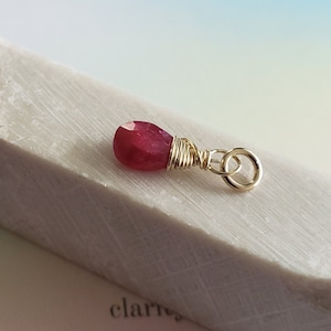 May include: A teardrop-shaped, faceted ruby gemstone pendant. The ruby is a deep red color and is wrapped in gold-colored wire. The pendant has a small loop at the top for attaching to a chain. The background is a light, neutral color.