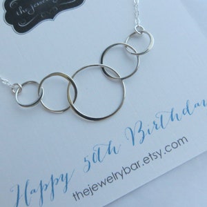 May include: A silver necklace with five interlocking circles, a popular gift for a 50th birthday. The necklace is on a white card with the text "Happy 50th Birthday!" and the website "thejewelrybar.etsy.com".