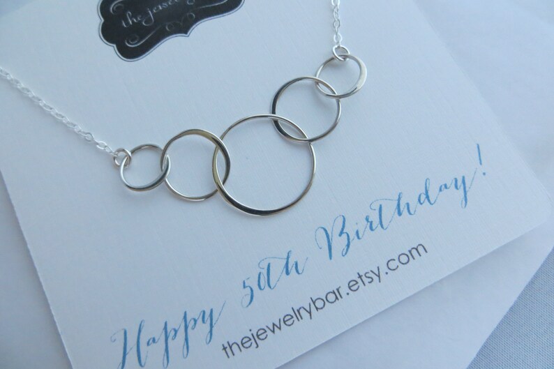 May include: A silver necklace with five interlocking circles, a popular gift for a 50th birthday. The necklace is on a white card with the text "Happy 50th Birthday!" and the website "thejewelrybar.etsy.com".