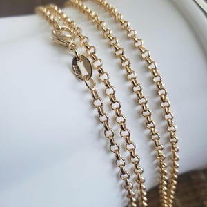 May include: Close-up of multiple gold-colored chain necklaces with a delicate, circular link design. The chains are draped over a white surface, with a clasp visible at the top. The chains have a shiny, reflective surface.