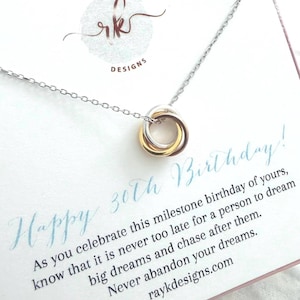 Puede incluir: Un collar con tres anillos entrelazados en tonos plata, oro y oro rosa en una cadena fina. La tarjeta de presentación dice: Happy 30th Birthday! As you celebrate this milestone birthday of yours, know that it is never too late for a person to dream big dreams and chase after them. Never abandon your dreams. raykdesigns.com