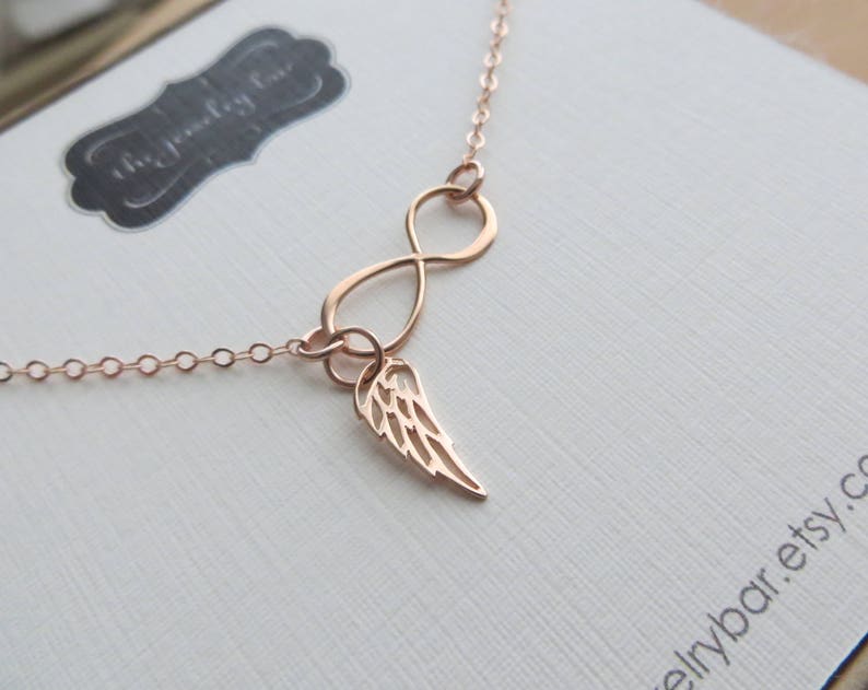 Pu&ograve; includere: Una collana a infinito in oro rosa con un delicato ciondolo ad ala d'angelo. La collana &egrave; su uno sfondo bianco.