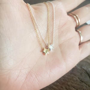 May include: Two delicate star pendant necklaces, one gold-toned and one silver-toned, rest on a palm. The necklaces feature small, five-pointed star charms suspended from fine chains. The background is a blurred view of a wooden surface.