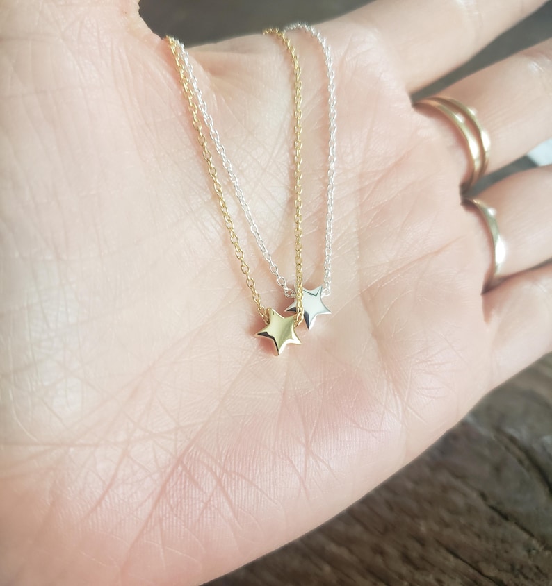 May include: Two delicate star pendant necklaces, one gold-toned and one silver-toned, rest on a palm. The necklaces feature small, five-pointed star charms suspended from fine chains. The background is a blurred view of a wooden surface.