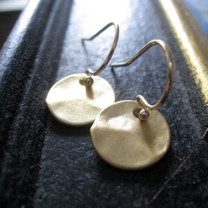 May include: A pair of gold-toned drop earrings featuring circular, hammered-texture discs suspended from thin, curved metal ear wires. Each disc is attached to the wire by a small, spherical bead, creating a simple, reflective, and metallic design.